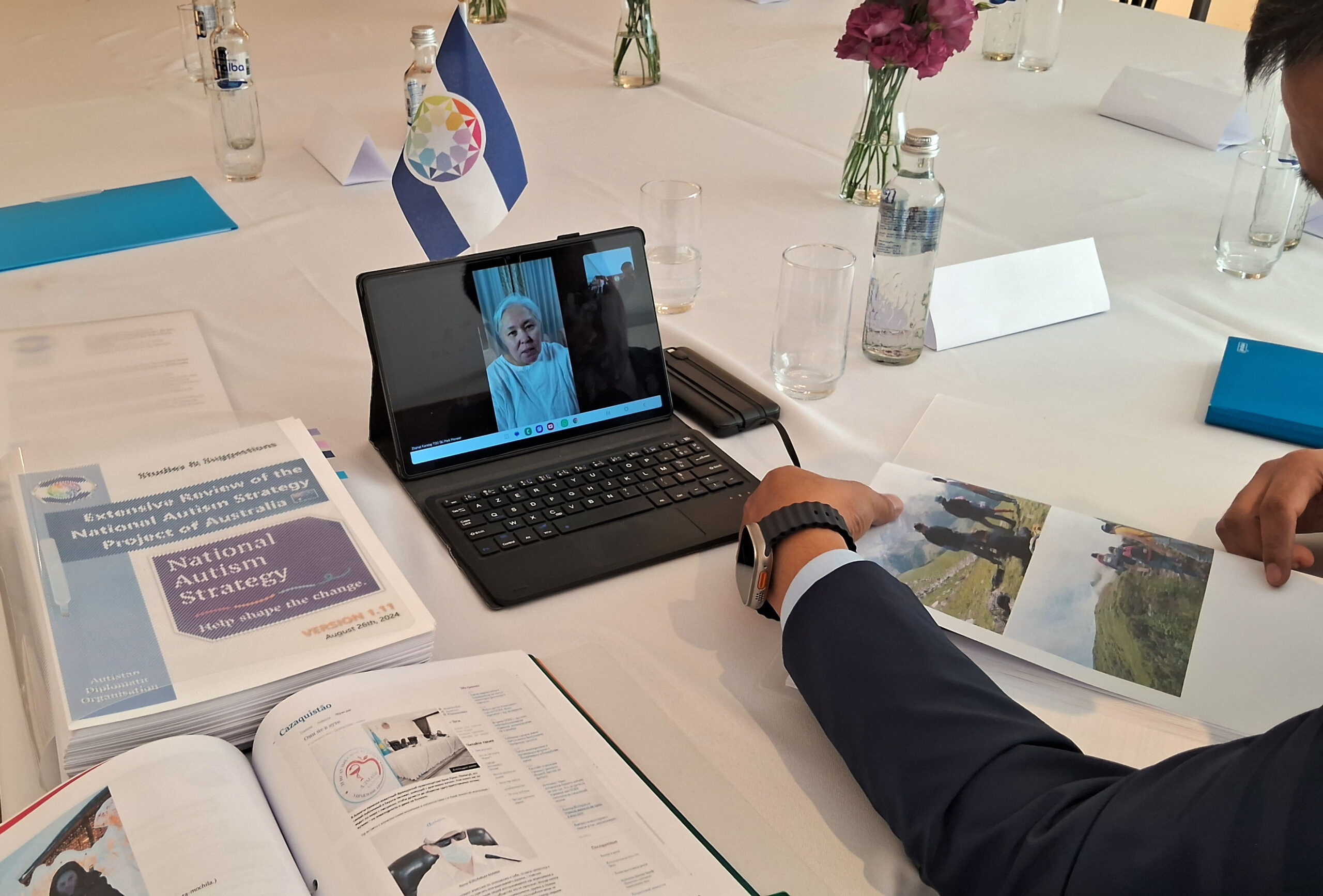 The videoconference with Zhanat Karatay, working documents on national autism strategies and the flag of the Diplomatic Organization of Autistan, while the Deputy Ambassador reads the printed documents.